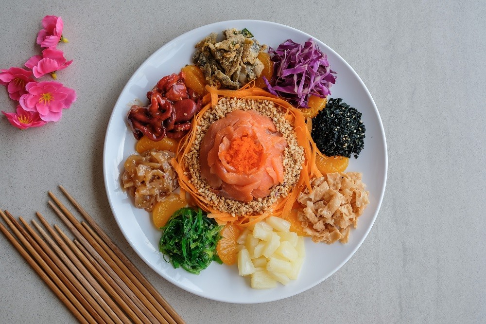 A large white plate of traditional yusheng featuring raw salmon slices, colorful vegetables, nuts, and fruits. A large white plate of traditional yusheng featuring raw salmon slices, colorful vegetables, nuts, and fruits.