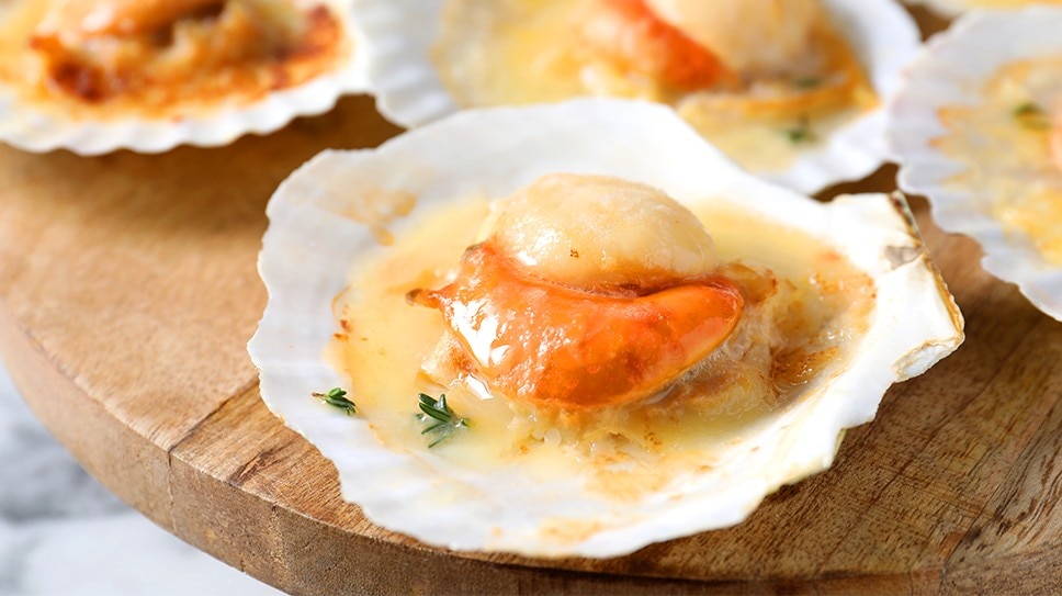 Freshly baked scallops on their shell on top of a wooden board.