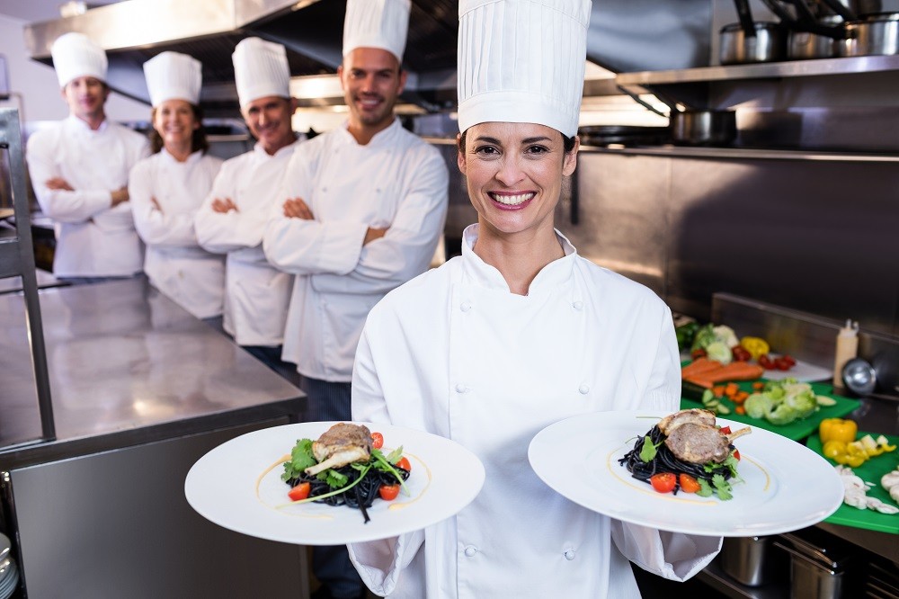 Female chef holding two plates of food while her colleagues flank her in the background Female chef holding two plates of food while her colleagues flank her in the background