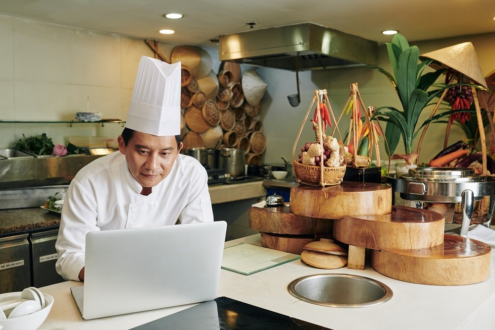 A female restaurant worker wearing a white chef’s jacket writing sales on a notebook and laptop during closing time A female restaurant worker wearing a white chef’s jacket writing sales on a notebook and laptop during closing time
