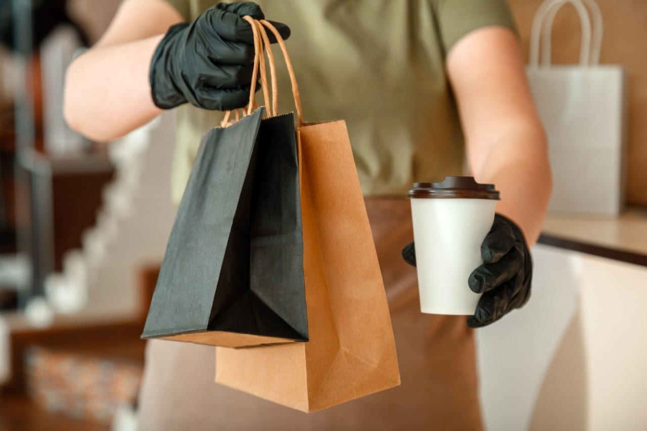 A waiter handing over two paper bags of takeout and a to-go cup. A waiter handing over two paper bags of takeout and a to-go cup.