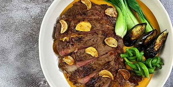 Ribeye steak and vegetables over kare-kare sauce in a white bowl.