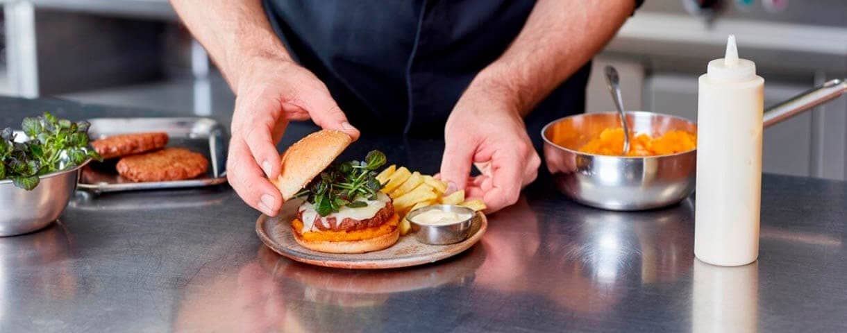 A cook assembling a burger with a side of fries on a plate.
