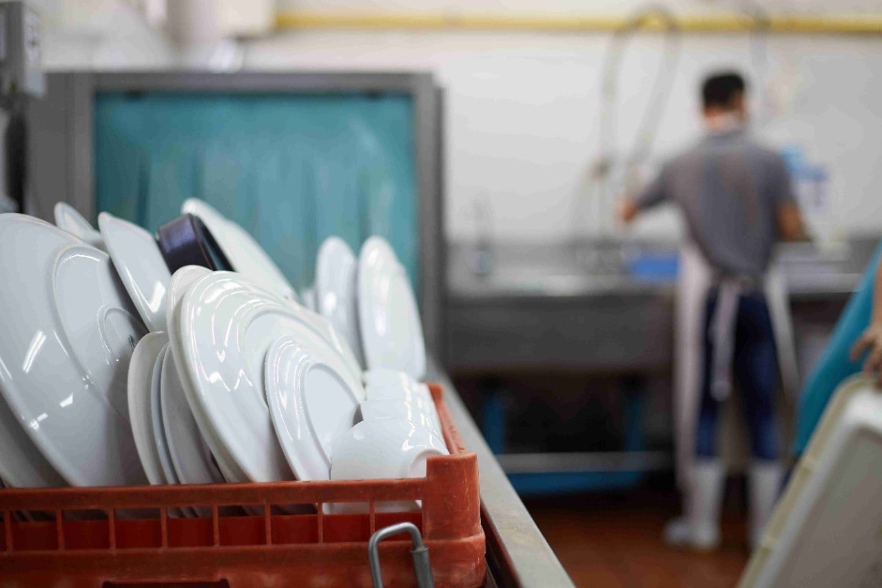 A crate filled with clean plates sits on the foreground while a lone dishwasher can be seen washing in the background A crate filled with clean plates sits on the foreground while a lone dishwasher can be seen washing in the background